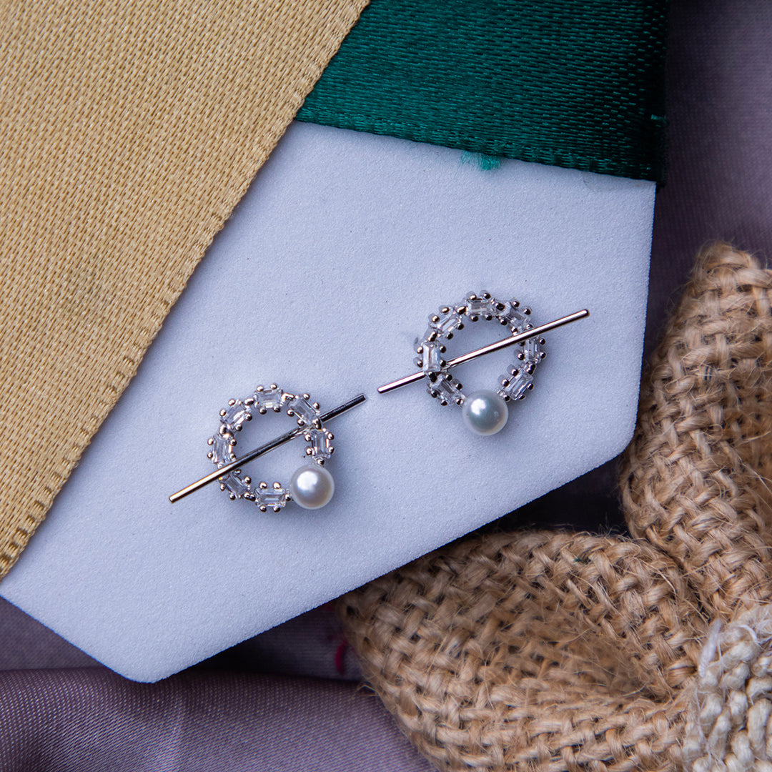 A pair of silver stud earrings with a geometric pattern, displayed on a white card, with a yellow textured background and a burlap fabric flower next to it.