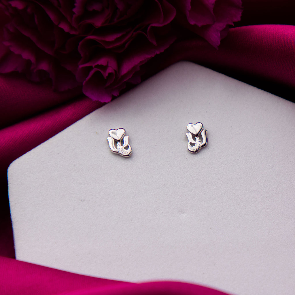 A pair of sterling silver heart-shaped stud earrings displayed on a white surface with a purple flower in the background.