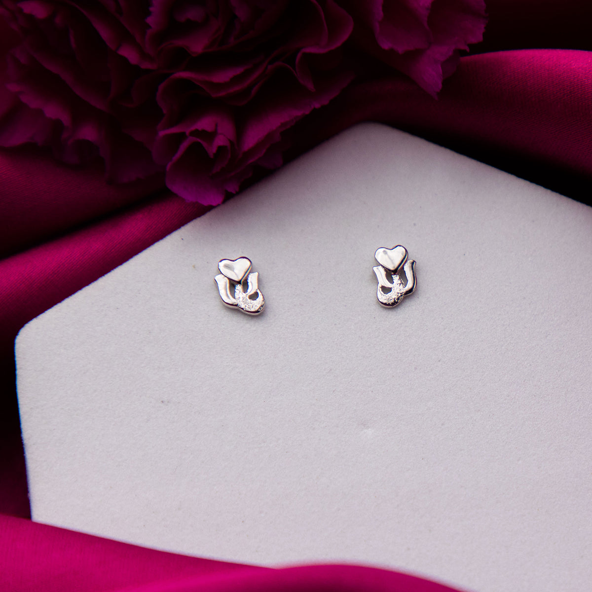 A pair of sterling silver heart-shaped stud earrings displayed on a white surface with a purple flower in the background.