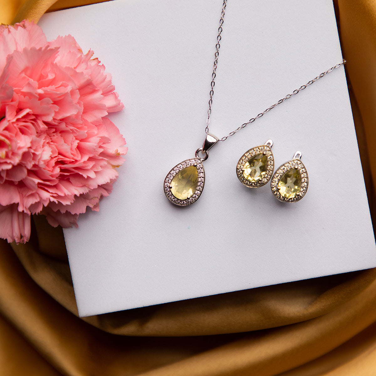 A sterling silver pendant set with a high shine finish and a pair of earrings, featuring a dazzling yellow stone pendant, displayed on a white surface with a pink flower and a gold-colored background.