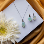 A sterling silver jade stone pendant set with a pair of earrings, displayed on a white surface with a yellow tint and a flower decoration behind it.