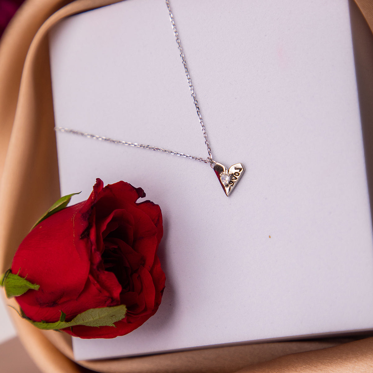 A silver pendant chain with a heart-shaped pendant featuring the word 'love' in the center, presented on a white cushion next to a red rose.