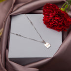 A silver pendant chain with a 'smile' motif on a grey background, presented in a box with a red flower beside it.
