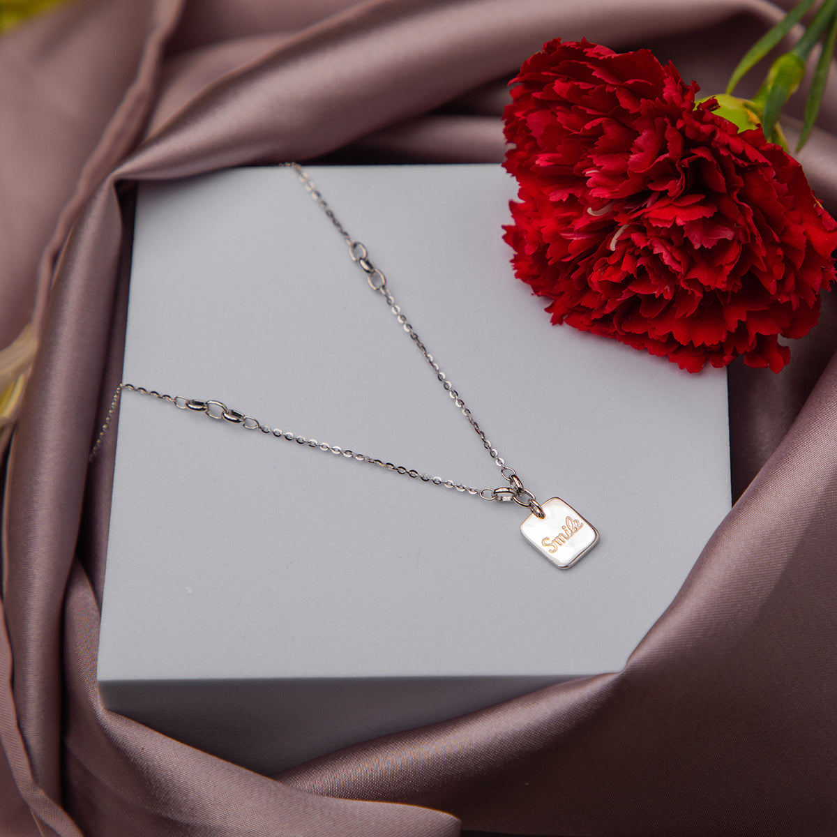 A silver pendant chain with a 'smile' motif on a grey background, presented in a box with a red flower beside it.