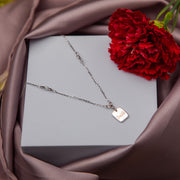 A silver pendant chain with a 'smile' motif on a grey background, presented in a box with a red flower beside it.