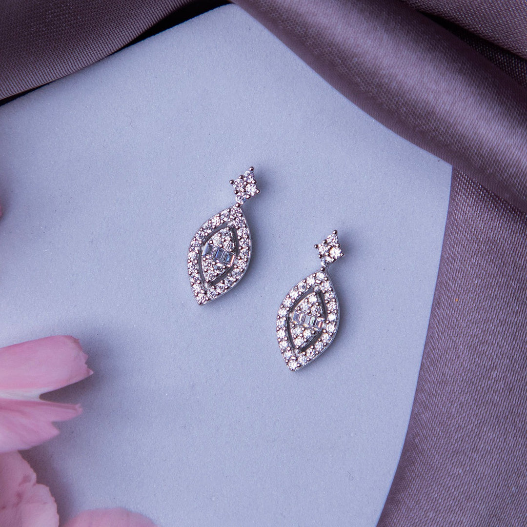A pair of silver leaf motif embellished earrings displayed on a surface with a grey fabric and pink flowers around.