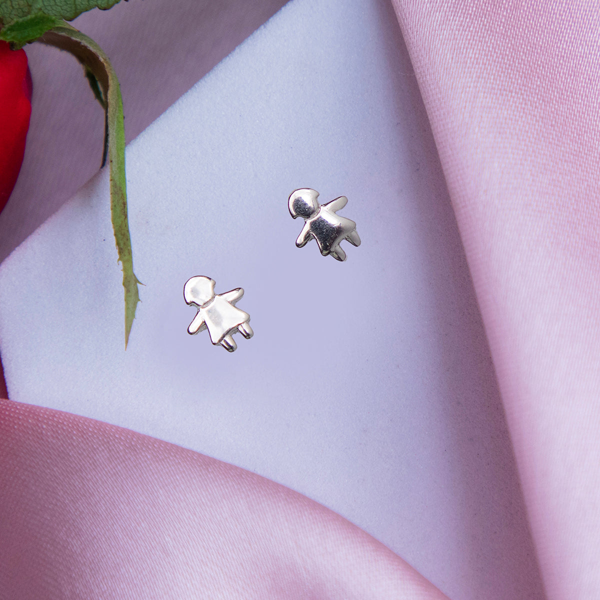 A pair of silver stud earrings designed to resemble a little girl, displayed on a pink fabric surface.