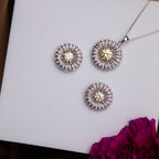 A sterling silver pendant featuring a dandelion seed motif accompanied by a pair of earrings with a similar design, displayed on a white surface with a purple flower beside them.