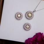 A sterling silver pendant featuring a dandelion seed motif accompanied by a pair of earrings with a similar design, displayed on a white surface with a purple flower beside them.