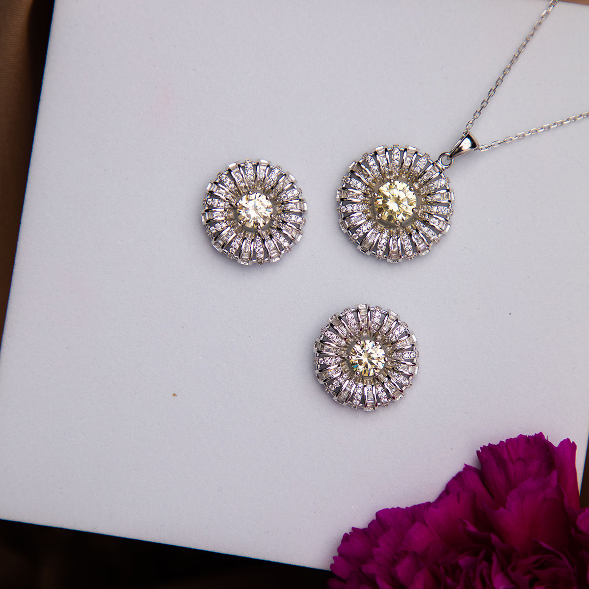 A sterling silver pendant featuring a dandelion seed motif accompanied by a pair of earrings with a similar design, displayed on a white surface with a purple flower beside them.