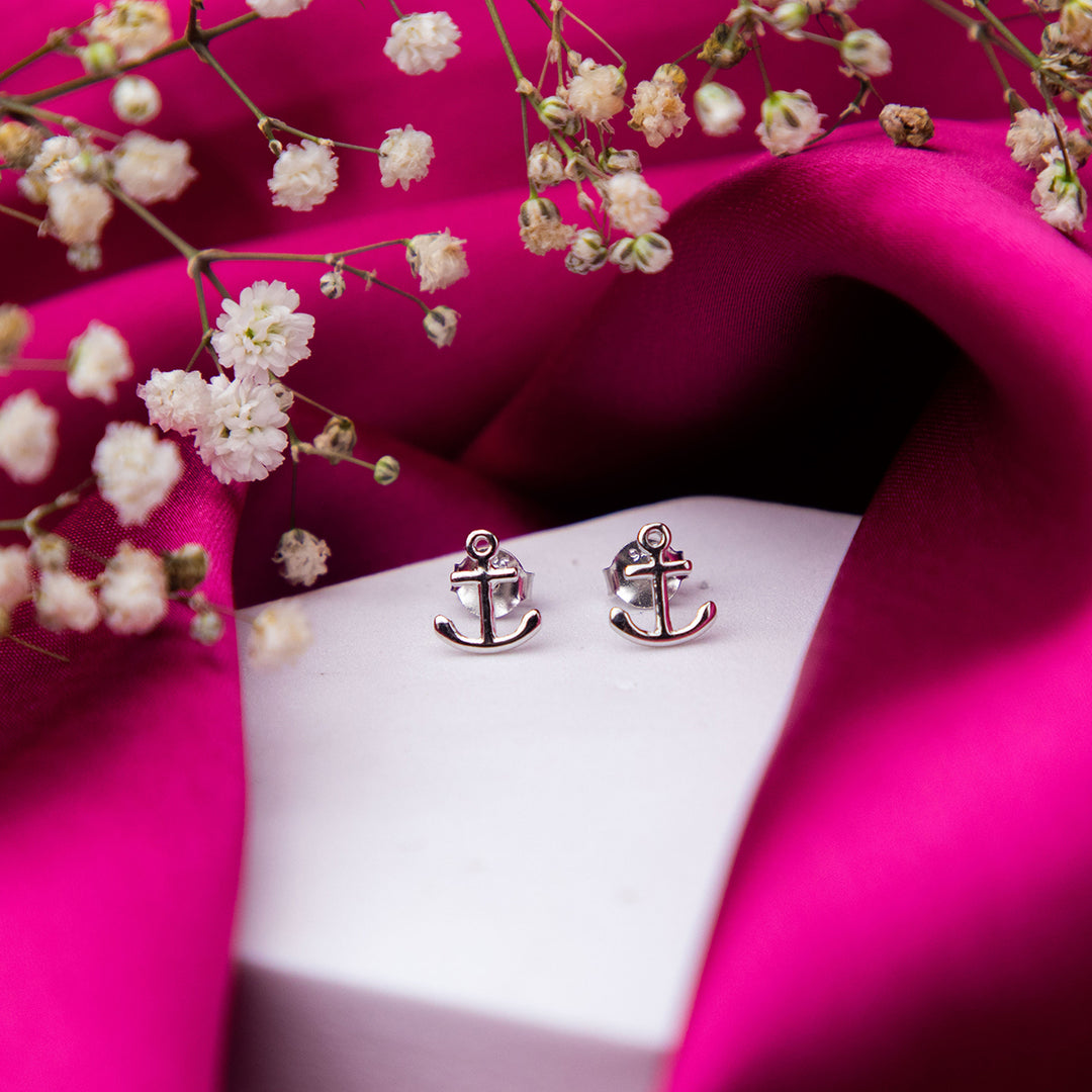 A pair of silver stud earrings designed to resemble an anchor, displayed on a pink fabric backdrop with decorative flowers around.