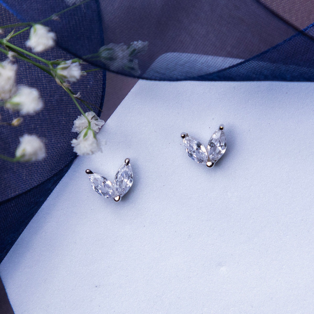 A pair of minimalistic white stone stud earrings placed on a white surface with a floral design in the background.