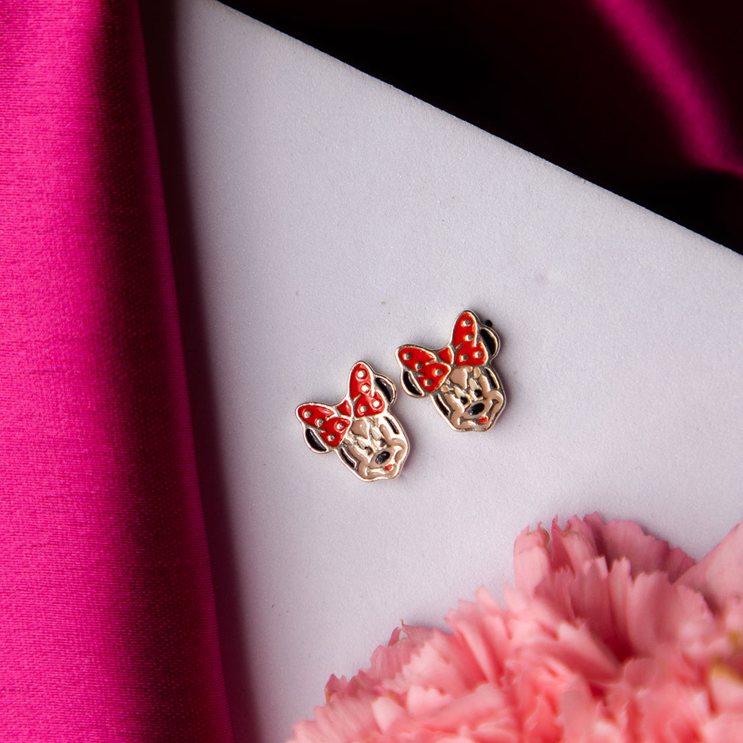 A pair of sterling silver Minnie Mouse fantasy stud earrings with red and black enamel accents, displayed on a white surface with a pink cloth and flower in the background.