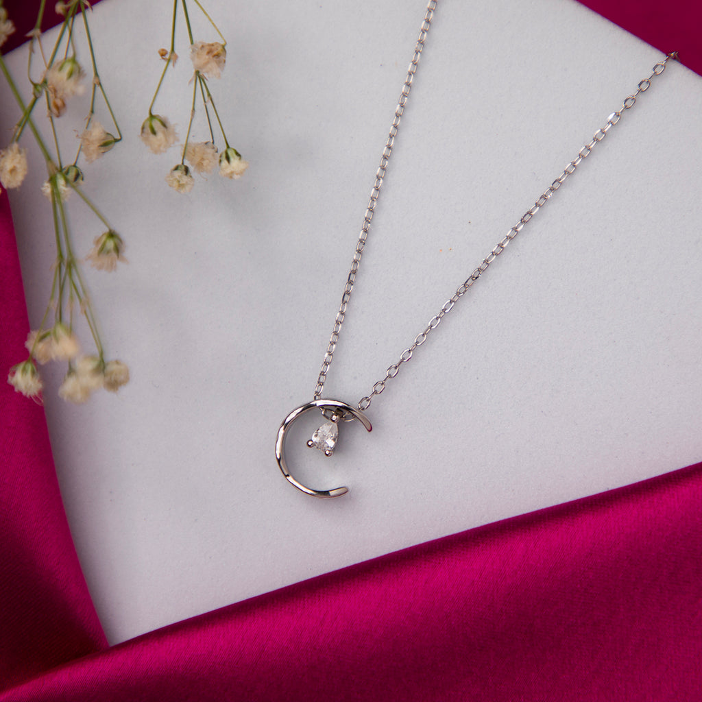 A silver pendant chain with a crescent moon design and a sparkling pear-cut white stone on a pink satin background with a flower in the background.