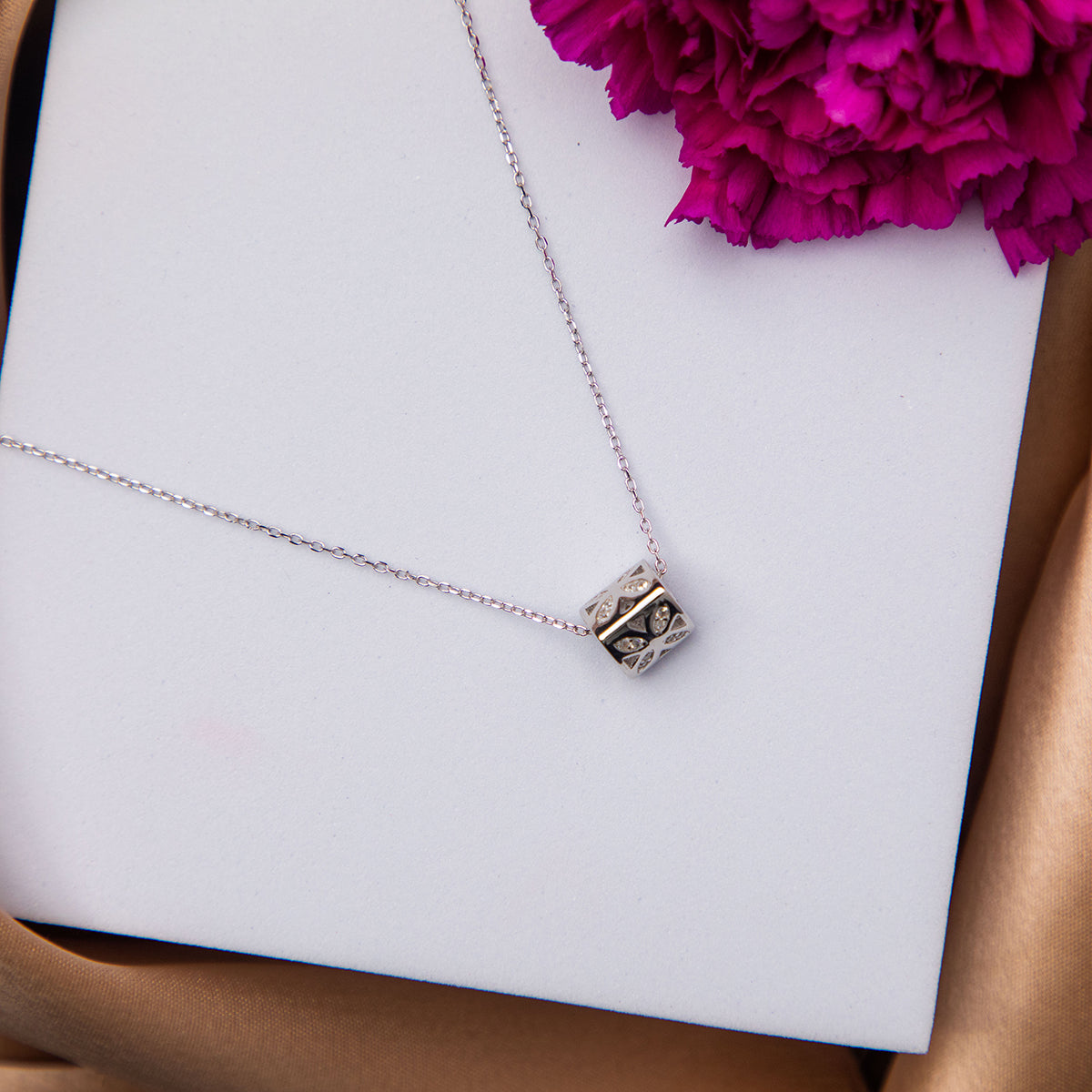 A sterling silver dainty pendant chain with a multifaceted geometric shape pendant placed on a white surface with a purple flower beside it.