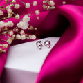 A pair of sterling silver mushroom-shaped stud earrings displayed on a white surface with a bright pink fabric and small white flowers in the background.