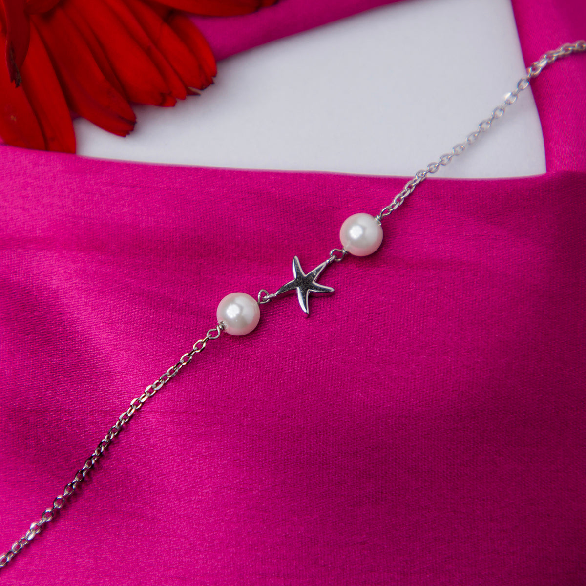 A silver bracelet featuring a starfish charm and pearl beads, displayed on a pink fabric surface.