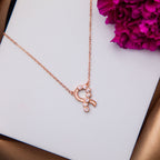 Rose gold-finish pendant chain necklace with a ribbon motif design, displayed on a white surface next to a purple flower.