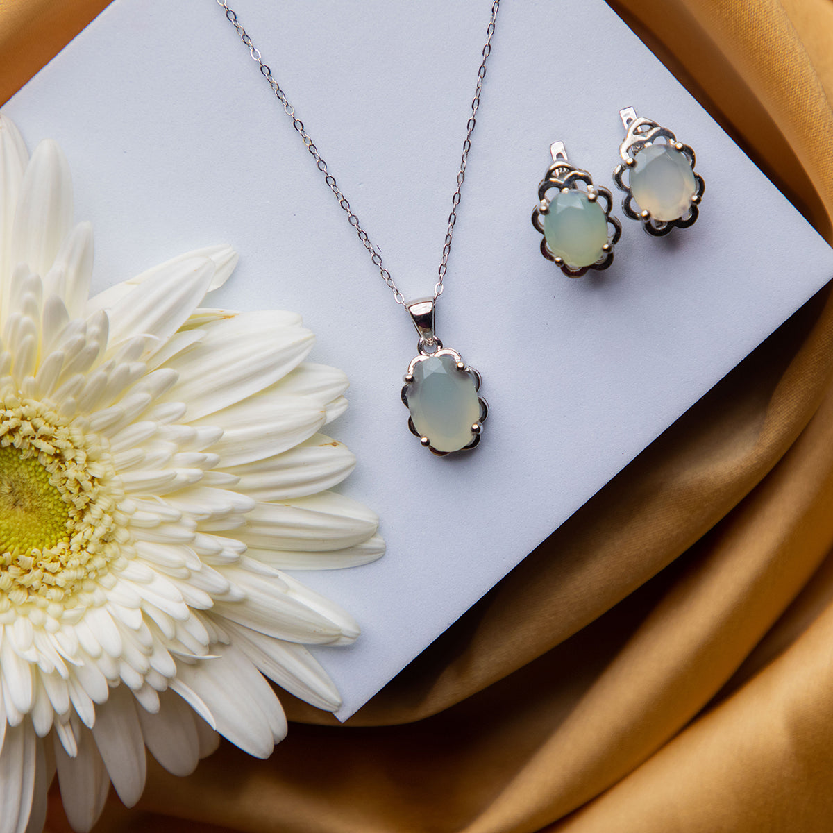 A sterling silver pendant set with a opalescent stone pendant and a pair of earrings, displayed on a white surface with a yellow and white flower background.