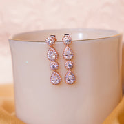 A pair of rose gold finished drop earrings with embellished gemstones displayed on a white cup.