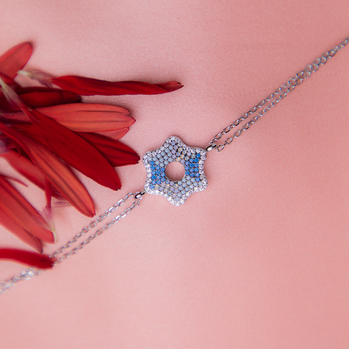 A sterling silver bracelet featuring a series of star-shaped charms with periwinkle blue gemstones, displayed on a pink background with a red flower scattered around.