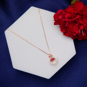 A rose gold-colored silver pendant chain with a heart-shaped pendant featuring a red moveable element, displayed on a white geometric-shaped stand with a red flower in the background.