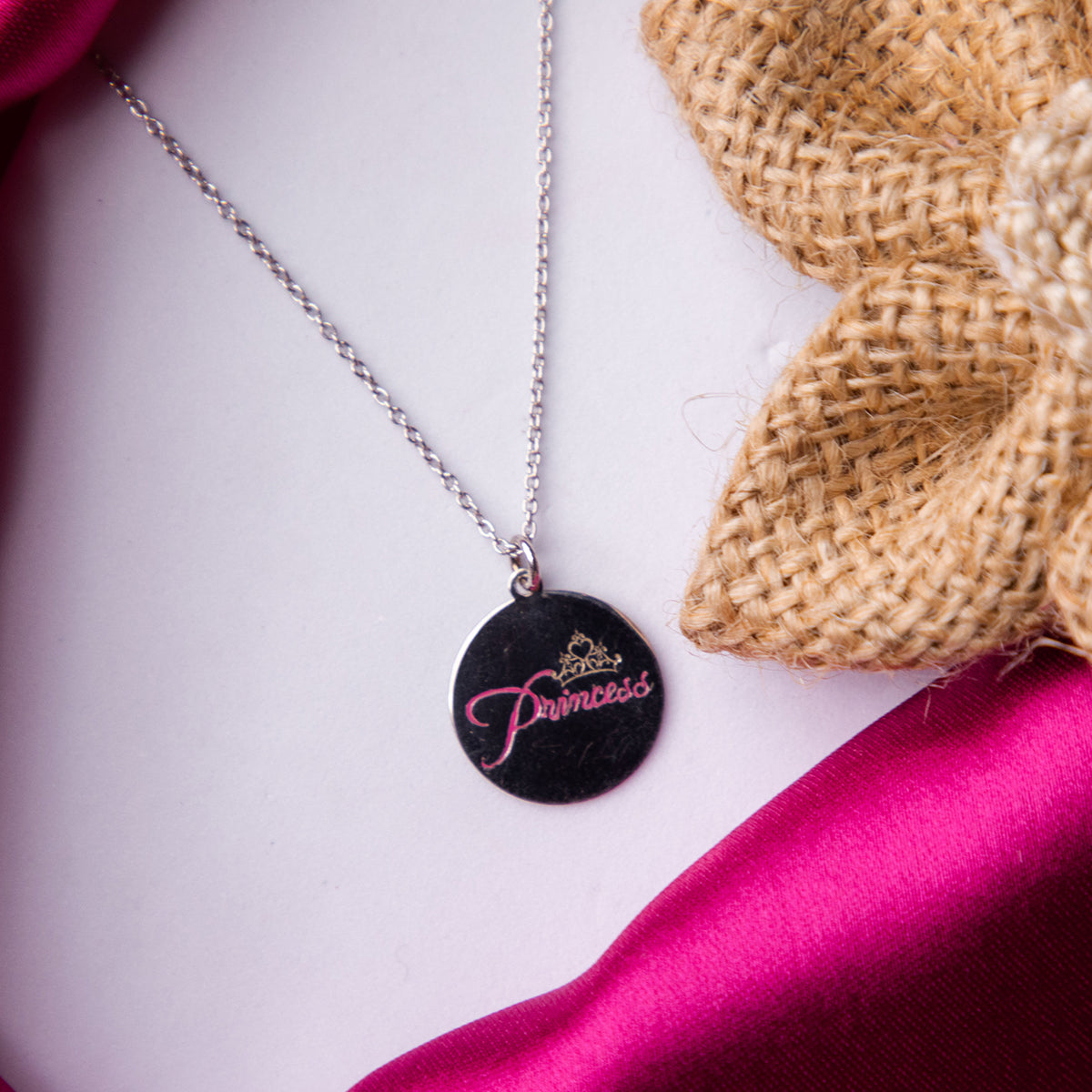 A sterling silver pendant chain with a round pendant engraved with the word 'Princess' presented on a pink fabric surface.