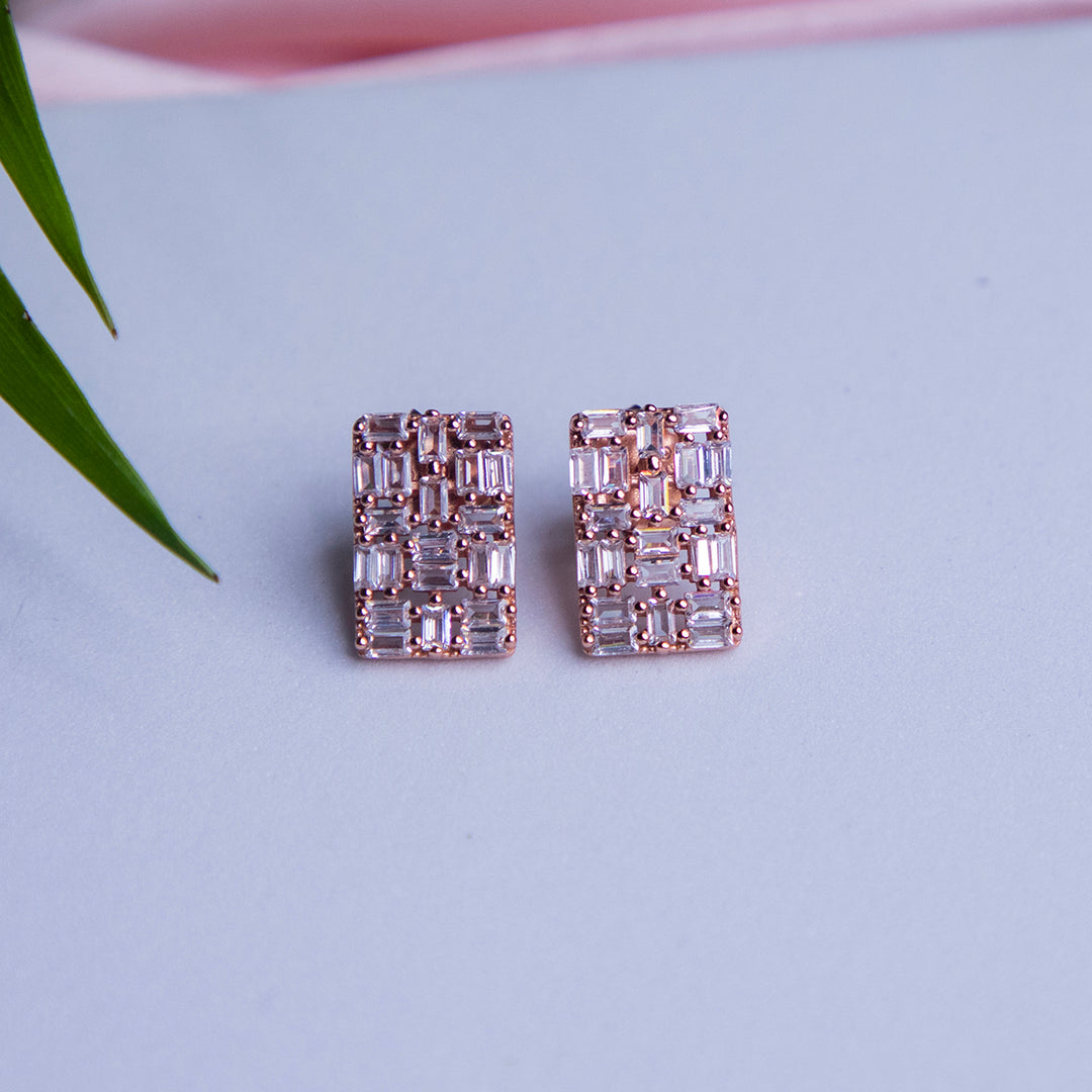 A pair of rose gold rectangle-shaped everyday silver stud earrings with sparkling embellishments, placed on a blue surface next to a green leaf.