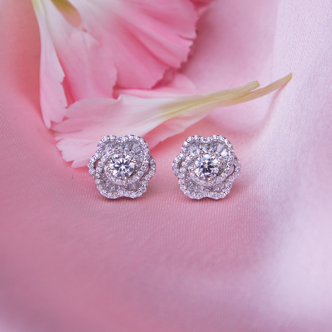 A pair of sterling silver rose-shaped stud earrings, displayed on a pink background with a petal motif.