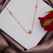 A rose gold colored pendant chain with a teardrop-shaped pendant, displayed on a white surface with a red rose and petals around it.