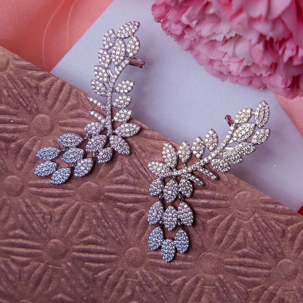 A pair of silver earcuffs designed to resemble an olive branch, displayed on a textured pink background with a flower for scale.