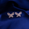 A pair of rose gold-colored butterfly-shaped dazzle stud earrings on a blue textured background.