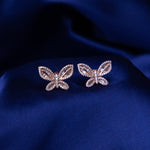 A pair of rose gold-colored butterfly-shaped dazzle stud earrings on a blue textured background.