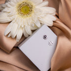 A pair of silver stud earrings with a silly eye emoji design, displayed on a soft surface with a white flower and a beige fabric background.