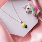 A sterling silver pendant chain featuring a yellow teddy bear with a green apple, displayed on a pink background.
