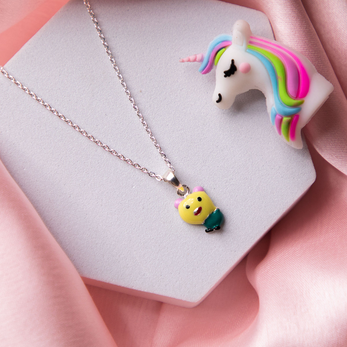 A sterling silver pendant chain featuring a yellow teddy bear with a green apple, displayed on a pink background.