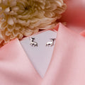 A pair of silver stag stud earrings displayed on a pink fabric surface with a white flower and a pale pink background.