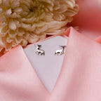 A pair of silver stag stud earrings displayed on a pink fabric surface with a white flower and a pale pink background.