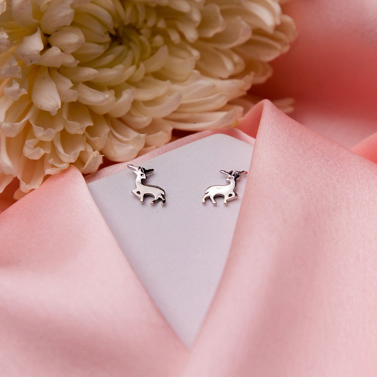 A pair of silver stag stud earrings displayed on a pink fabric surface with a white flower and a pale pink background.