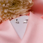 A pair of silver stag stud earrings displayed on a pink fabric surface with a white flower and a pale pink background.