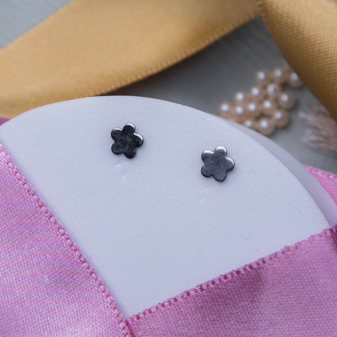 A pair of silver stud earrings with a flower motif, displayed on a pink textured surface.