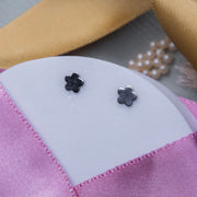 A pair of silver stud earrings with a flower motif, displayed on a pink textured surface.