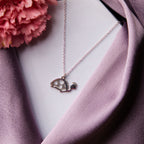 A silver pendant chain in the shape of a tortoise displayed on a white base wearing a purple fabric and a pink flower is visible in the background.