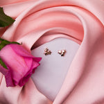 A pair of rose gold finish stud earrings with a star cluster design, displayed on a surface with a pink fabric and a rose flower.