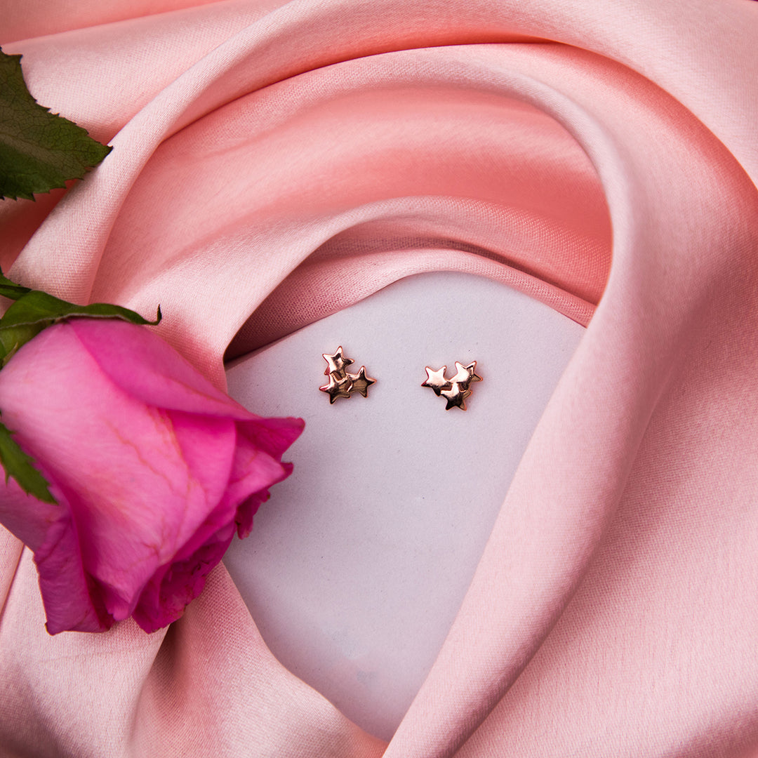 A pair of rose gold finish stud earrings with a star cluster design, displayed on a surface with a pink fabric and a rose flower.