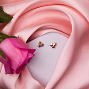 A pair of rose gold finish stud earrings with a star cluster design, displayed on a surface with a pink fabric and a rose flower.