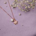 A rose gold-colored silver pendant chain with a starfish motif and a pearl, displayed on a purple background with some scattered flower petals.