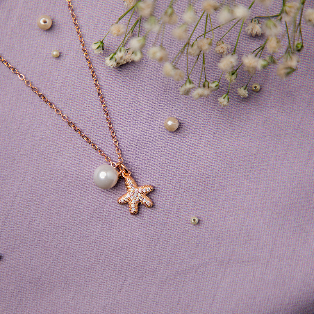 A rose gold-colored silver pendant chain with a starfish motif and a pearl, displayed on a purple background with some scattered flower petals.