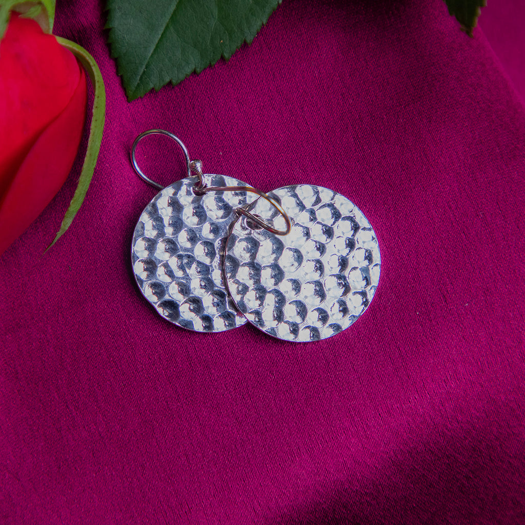 A pair of round silver earrings with a tribal hammered pattern, displayed on a pink background with a red rose and green leaves.