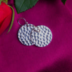 A pair of round silver earrings with a tribal hammered pattern, displayed on a pink background with a red rose and green leaves.